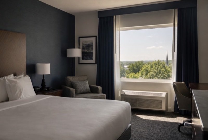 A calm hotel guest room with a made white bed, an armchair, a desk lamp and treetops through a large window, typical of the hotel industry